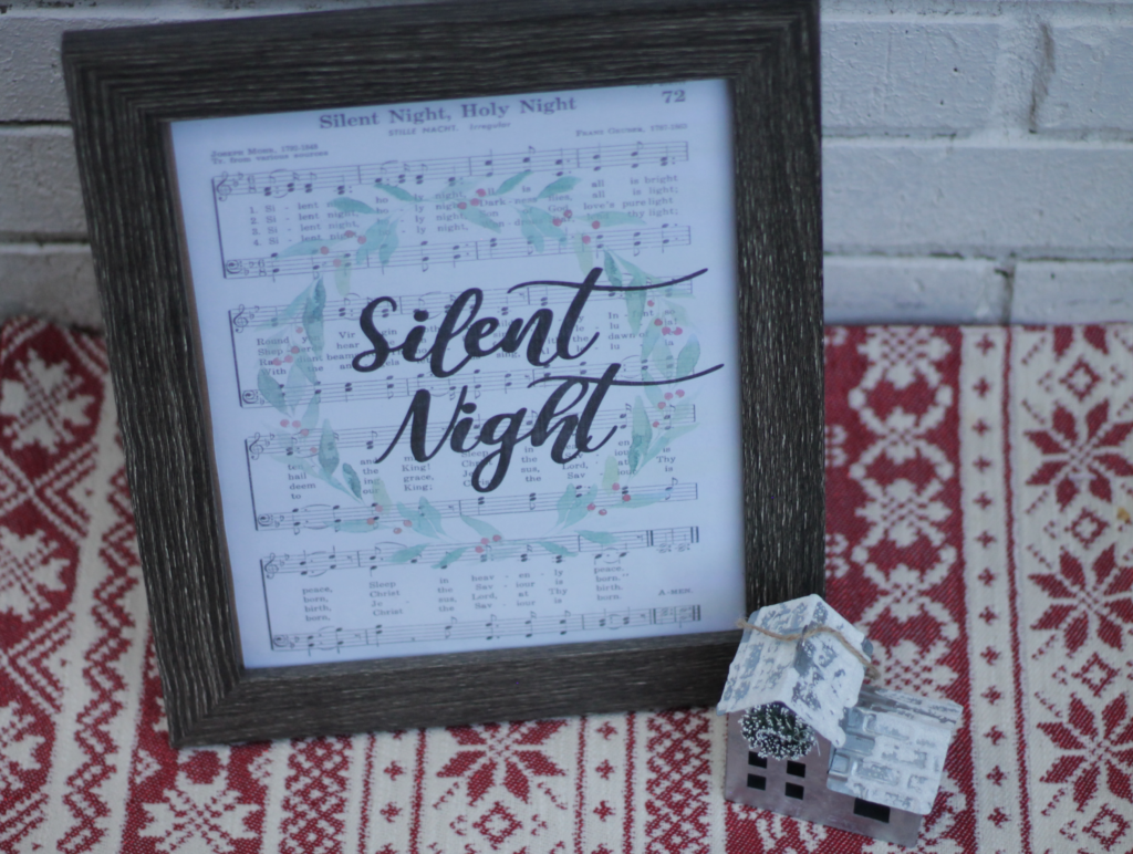 wooden frame in front of white brick wall with Silent night sheet music inside displaying a Christmas wreath and script text of silent night in the center, beside a metal house decor on top of red and cream Christmas designed fabric 