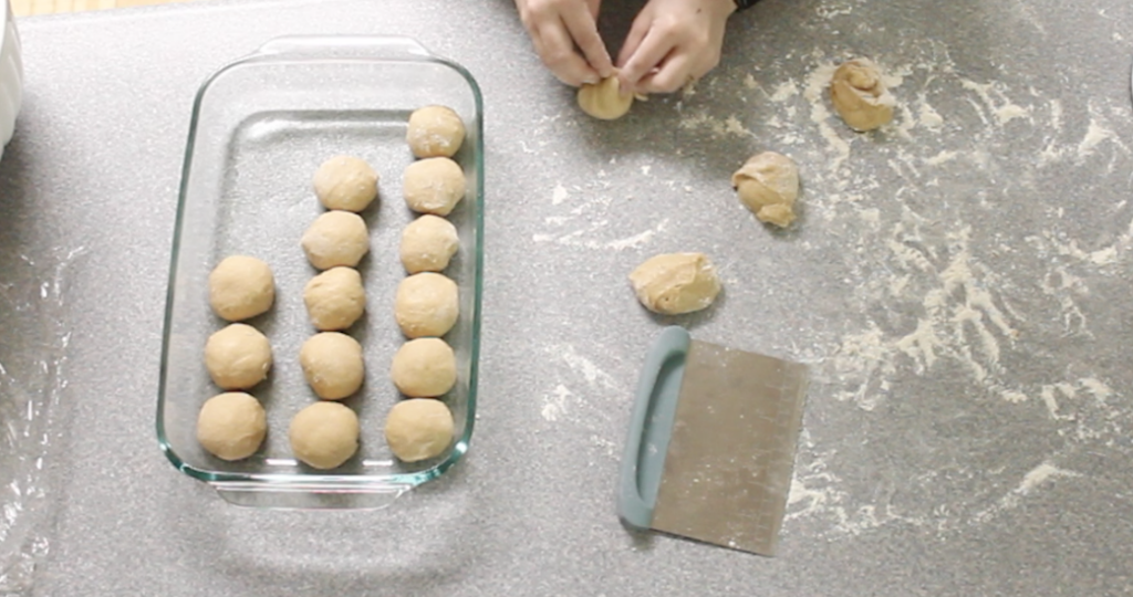 woman shaping dough into rolls with bench scraper on counter and shaped rolls in 9x13 glass pan 