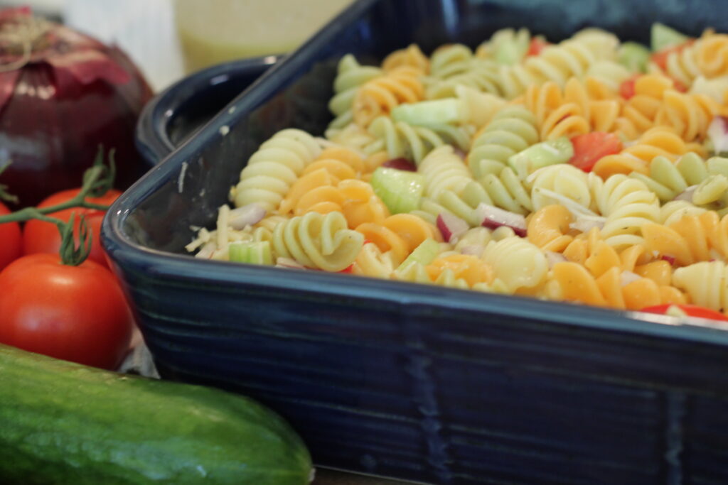 pasta salad in blue dish with onion, tomatoes, and cucumber on the left.